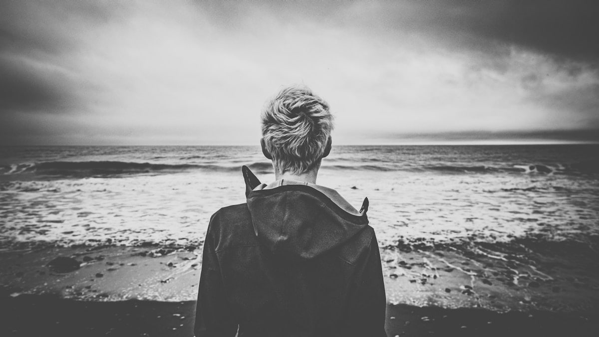 grayscale photo of woman in black coat standing on beach